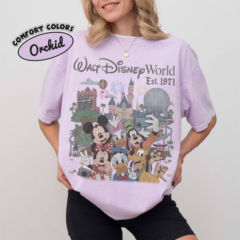May include: An orchid-colored t-shirt featuring a graphic print of Walt Disney World Est. 1971. The design includes illustrations of Mickey Mouse, Minnie Mouse, Donald Duck, Daisy Duck, Goofy, Pluto, Chip, and Dale in front of park landmarks.