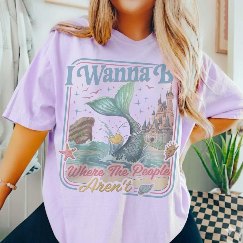 May include: A light purple t-shirt with a graphic design featuring a mermaid tail, a castle, and the text "I Wanna B Where The People Aren't". The design includes a starfish, seashells, and a duck.