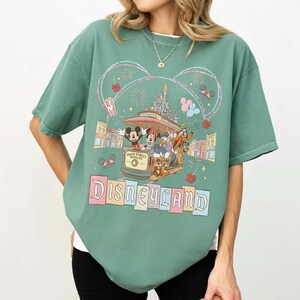 May include: Green t-shirt featuring a Disneyland design. The graphic includes Mickey Mouse, Minnie Mouse, Donald Duck, and Goofy riding a Main Street Trolley. The word "Disneyland" is displayed in colorful block letters.