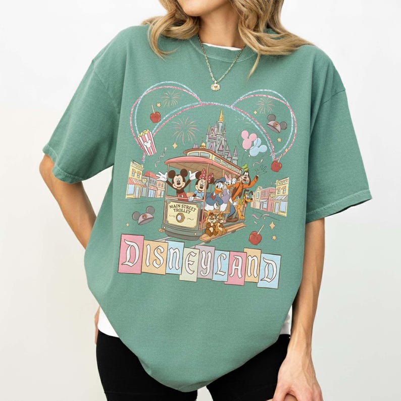 May include: Green t-shirt featuring a Disneyland design. The graphic includes Mickey Mouse, Minnie Mouse, Donald Duck, and Goofy riding a Main Street Trolley. The word "Disneyland" is displayed in colorful block letters.