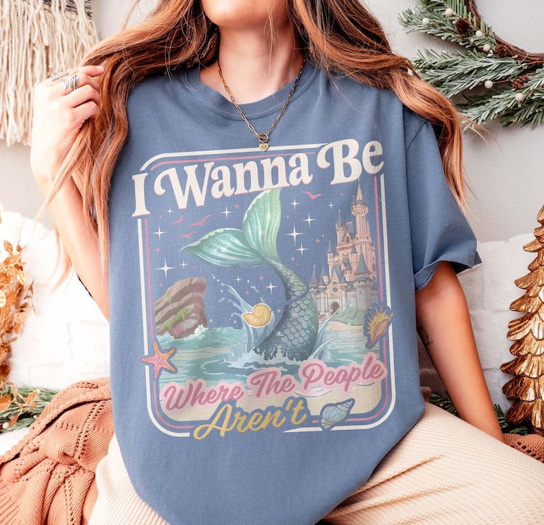 May include: Blue t-shirt with a mermaid graphic and the text "I Wanna Be Where The People Aren't". The design features a mermaid, castle, and ocean elements. The shirt is made of a soft material.