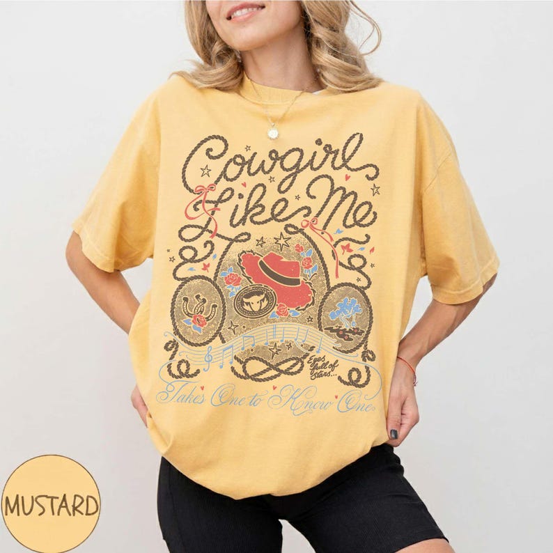 May include: A mustard yellow short-sleeve t-shirt featuring a graphic print with the text "Cowgirl Like Me," "Eyes full of stars," and "Takes One to Know One." The design includes a red cowboy hat, rope borders, musical notes, and stars.