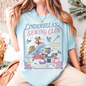 May include: Light blue t-shirt featuring the text "Cinderella's Sewing Club" with a graphic of Cinderella's glass slipper, mice, and sewing notions. The design includes the words "Bibbidi Bobbidi Boo" and "Est. 1950".