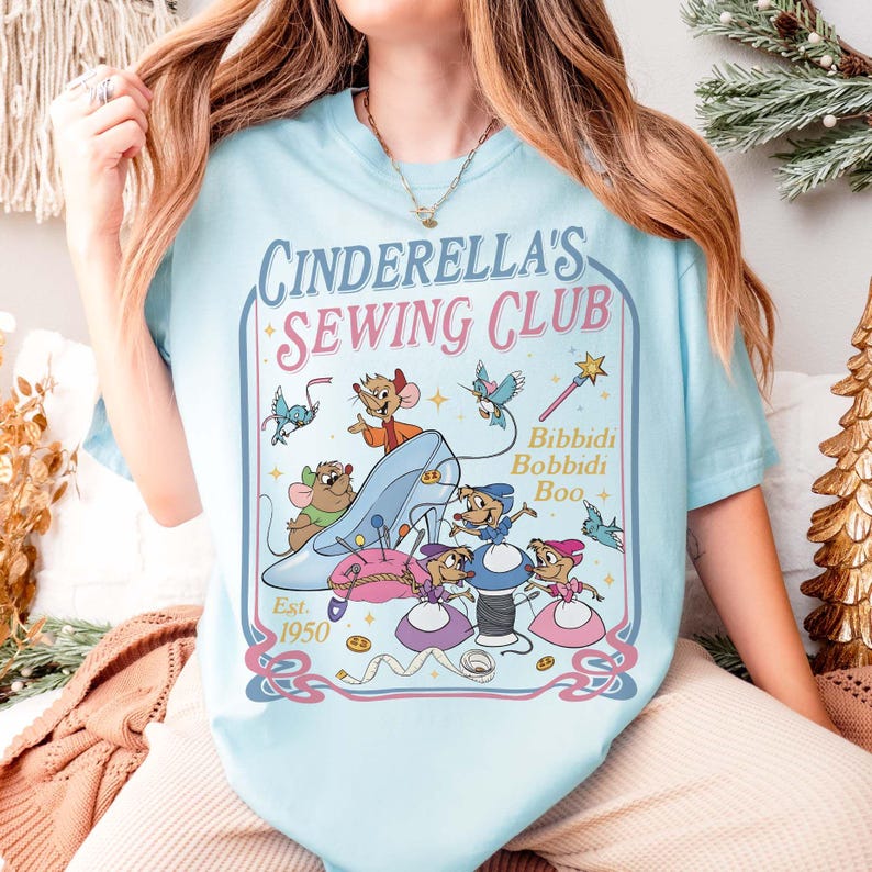 May include: Light blue t-shirt featuring the text "Cinderella's Sewing Club" with a graphic of Cinderella's glass slipper, mice, and sewing notions. The design includes the words "Bibbidi Bobbidi Boo" and "Est. 1950".