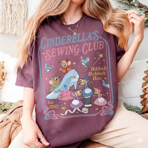 May include: A maroon t-shirt featuring a graphic of Cinderella's Sewing Club. The design includes a blue glass slipper, mice, and the text "Cinderella's Sewing Club" and "Bibbidi Bobbidi Boo". The shirt is dated "Est. 1950".