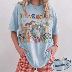 May include: A light blue short-sleeve t-shirt featuring a graphic print of various animated characters from the Toy Story franchise. The text "YOU'VE GOT A FRIEND IN ME" is printed in colorful, blocky letters above and below the character group.