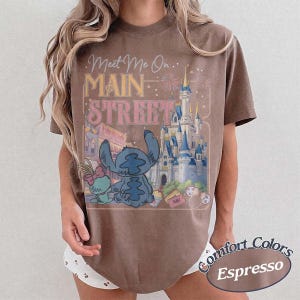 May include: Espresso-colored t-shirt featuring a graphic of Stitch and Angel in front of a castle with the text "Meet Me On Main Street." The shirt is from Comfort Colors.