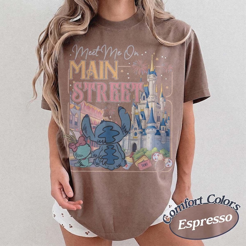 May include: Espresso-colored t-shirt featuring a graphic of Stitch and Angel in front of a castle with the text "Meet Me On Main Street." The shirt is from Comfort Colors.