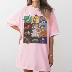 May include: A light pink short-sleeved t-shirt featuring a grid of nine rectangular panels. Each panel displays a different animated character, including a green dinosaur, a cowboy, a space ranger, a dog, a woman in a dress, a cowgirl, two potatoes, a horse, aliens, and a white spork character.