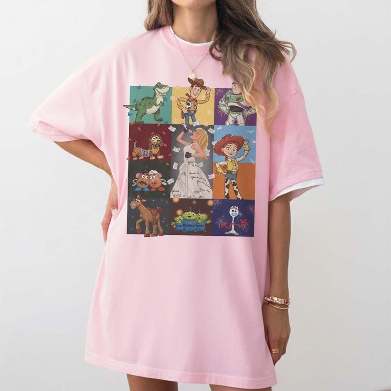 May include: A light pink short-sleeved t-shirt featuring a grid of nine rectangular panels. Each panel displays a different animated character, including a green dinosaur, a cowboy, a space ranger, a dog, a woman in a dress, a cowgirl, two potatoes, a horse, aliens, and a white spork character.