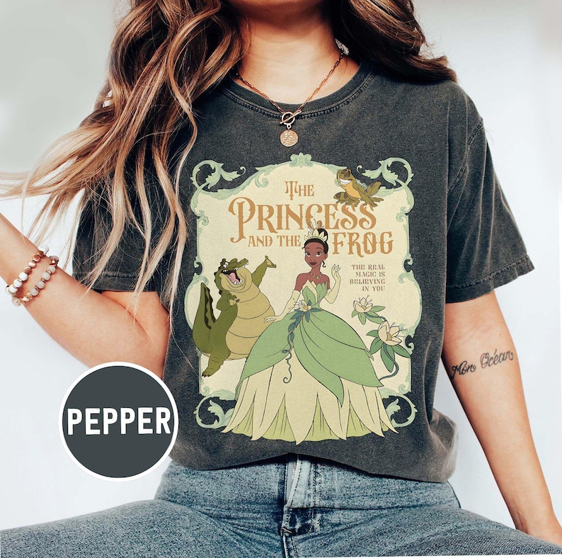 May include: Dark gray t-shirt featuring a graphic from "The Princess and the Frog." The design includes the title, characters, and the phrase "The Real Magic Is Believing In You." The shirt has a vintage, worn look.