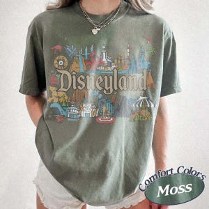 May include: Olive green t-shirt with a Disneyland Resort graphic. The graphic features colorful illustrations of various park attractions and the word "Disneyland" in a vintage font. The shirt also has the text "Comfort Colors Moss" on the bottom right.