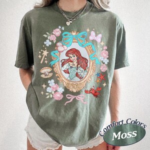 Puede incluir: Camiseta verde oliva con una ilustraci&oacute;n enmarcada de una mujer pelirroja con un vestido azul, rodeada de flores rosas, lazos azules y un cangrejo rojo. La camiseta tiene las palabras "Comfort Colors Moss" impresas.