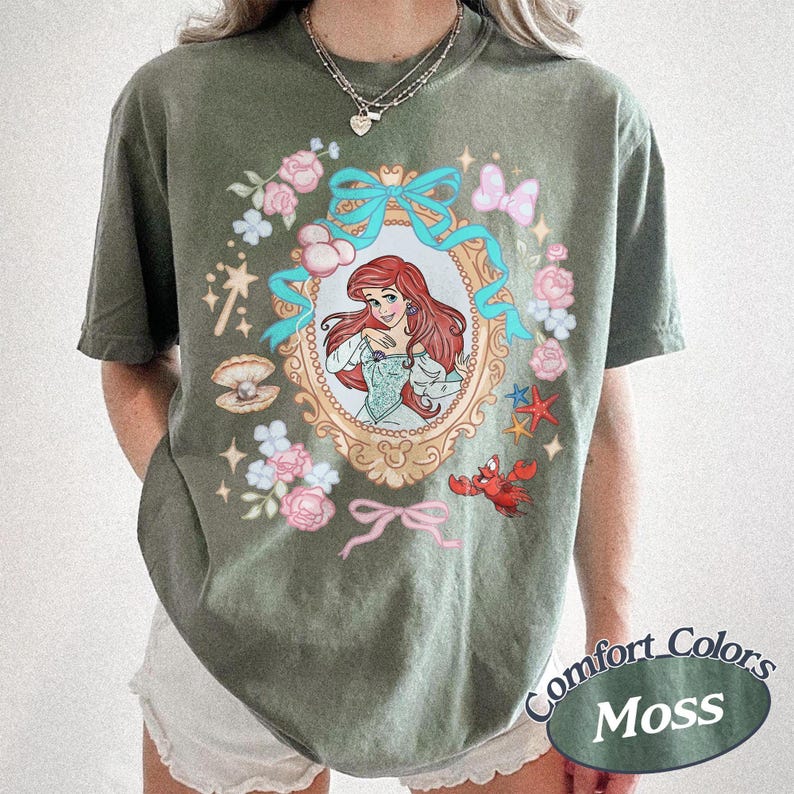 Puede incluir: Camiseta verde oliva con una ilustraci&oacute;n enmarcada de una mujer pelirroja con un vestido azul, rodeada de flores rosas, lazos azules y un cangrejo rojo. La camiseta tiene las palabras "Comfort Colors Moss" impresas.