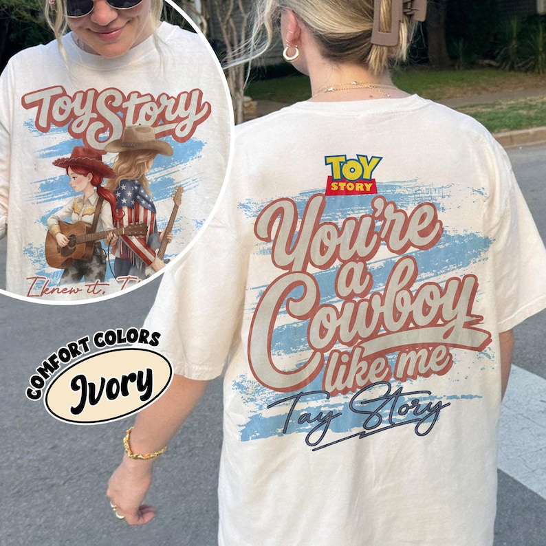 May include: An ivory t-shirt featuring a graphic print on the back with the text Toy Story, You're a Cowboy like me, and Tay Story. The design includes a blue brushstroke background and an illustration of two figures in cowboy hats playing guitars.
