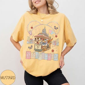 May include: Mustard yellow t-shirt featuring a Disneyland graphic. The design includes a trolley with Mickey Mouse, Minnie Mouse, Donald Duck, and other characters. The word "Disneyland" is displayed in pastel colors.