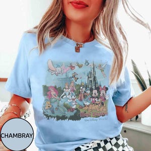 K&ouml;nnte beinhalten: Hellblaues T-Shirt mit einem farbenfrohen Aufdruck von Disney-Figuren, darunter Micky und Minnie Maus, Arielle und Dumbo, mit dem Text "Magic Kingdom Walt Disney World". Das Shirt hat kurze &Auml;rmel und einen Rundhalsausschnitt.