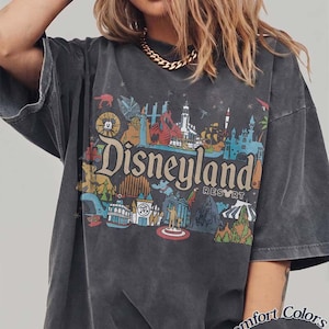 May include: Dark gray t-shirt with a colorful Disneyland Resort graphic. The design features various park icons and the Disneyland logo. The shirt has short sleeves and a relaxed fit. The text "Comfort Colors Pepper" is printed on the bottom right.