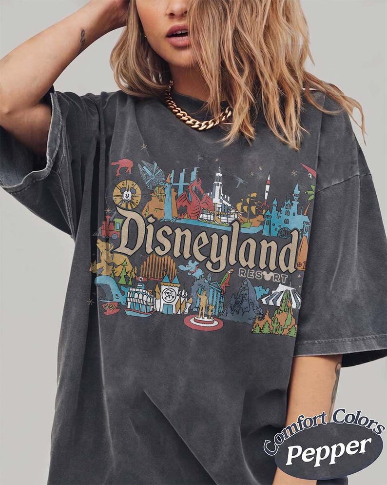 May include: Dark gray t-shirt with a colorful Disneyland Resort graphic. The design features various park icons and the Disneyland logo. The shirt has short sleeves and a relaxed fit. The text "Comfort Colors Pepper" is printed on the bottom right.