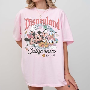 May include: A light pink t-shirt featuring a Disneyland graphic with Mickey Mouse, Minnie Mouse, Goofy, Donald Duck, and Pluto. The graphic includes the words "Disneyland California" and "Est 1955".