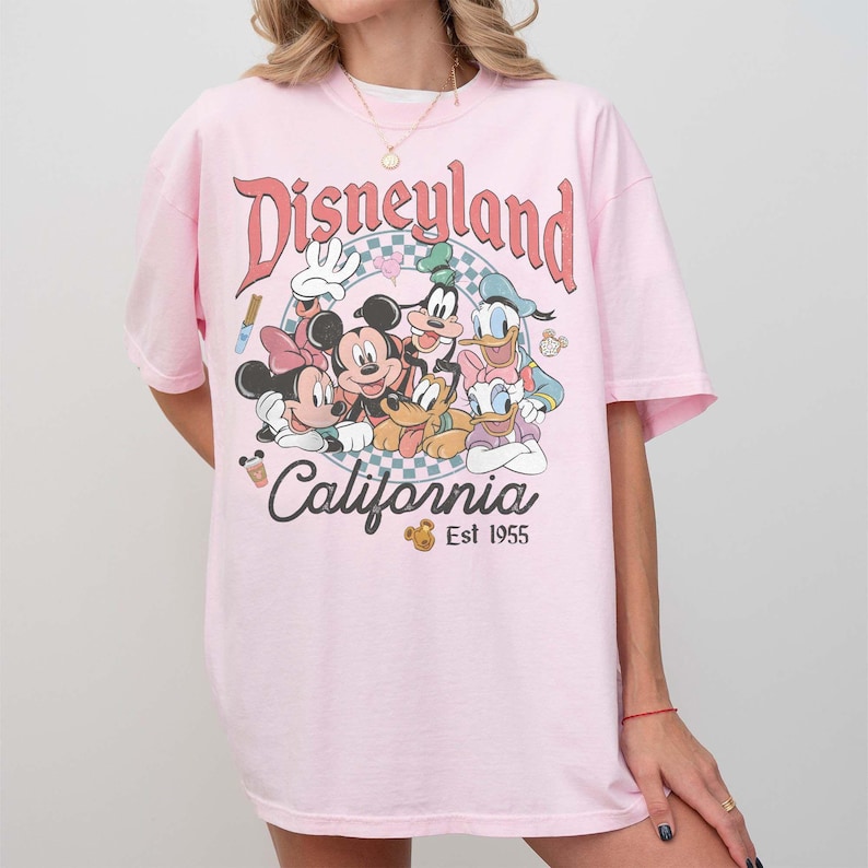 May include: A light pink t-shirt featuring a Disneyland graphic with Mickey Mouse, Minnie Mouse, Goofy, Donald Duck, and Pluto. The graphic includes the words "Disneyland California" and "Est 1955".