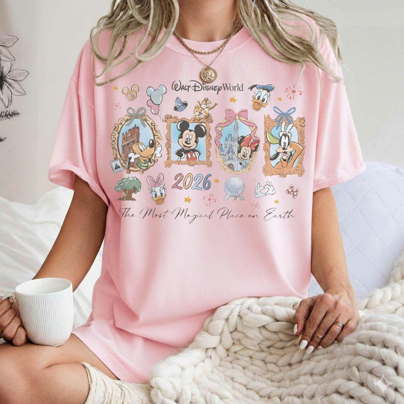 May include: Pink t-shirt featuring framed Disney characters, including Mickey and Minnie Mouse, Donald Duck, and Goofy. The shirt has the text "Walt Disney World" and "2026". The phrase "The Most Magical Place on Earth" is also printed on the shirt.