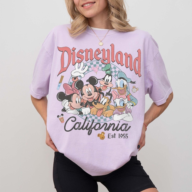 May include: A light purple t-shirt featuring a graphic of Disney characters including Mickey Mouse, Minnie Mouse, Donald Duck, and Goofy. The shirt has the words "Disneyland California" and "Est 1955" printed on it.