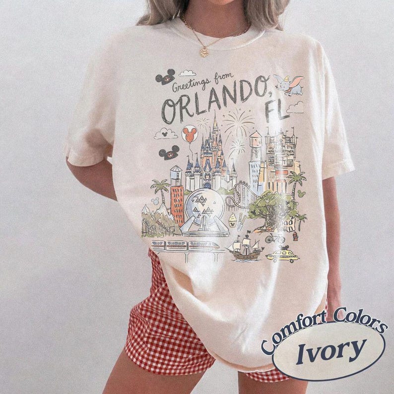 K&ouml;nnte beinhalten: Elfenbeinfarbenes T-Shirt mit einer Grafik von Wahrzeichen von Orlando, FL, darunter ein Schloss und Feuerwerk. Das Shirt tr&auml;gt den Text "Greetings from Orlando, FL" und "Comfort Colors Ivory".