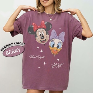 Pu&ograve; includere: Una t-shirt color bacca con una stampa grafica di Minnie Mouse e Daisy Duck. Il design include il testo Minnie Mouse e Daisy Duck con piccoli accenti a forma di stella. Un'etichetta circolare sul lato sinistro riporta Comfort Colors Berry.