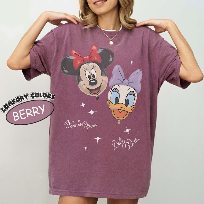 Pu&ograve; includere: Una t-shirt color bacca con una stampa grafica di Minnie Mouse e Daisy Duck. Il design include il testo Minnie Mouse e Daisy Duck con piccoli accenti a forma di stella. Un'etichetta circolare sul lato sinistro riporta Comfort Colors Berry.
