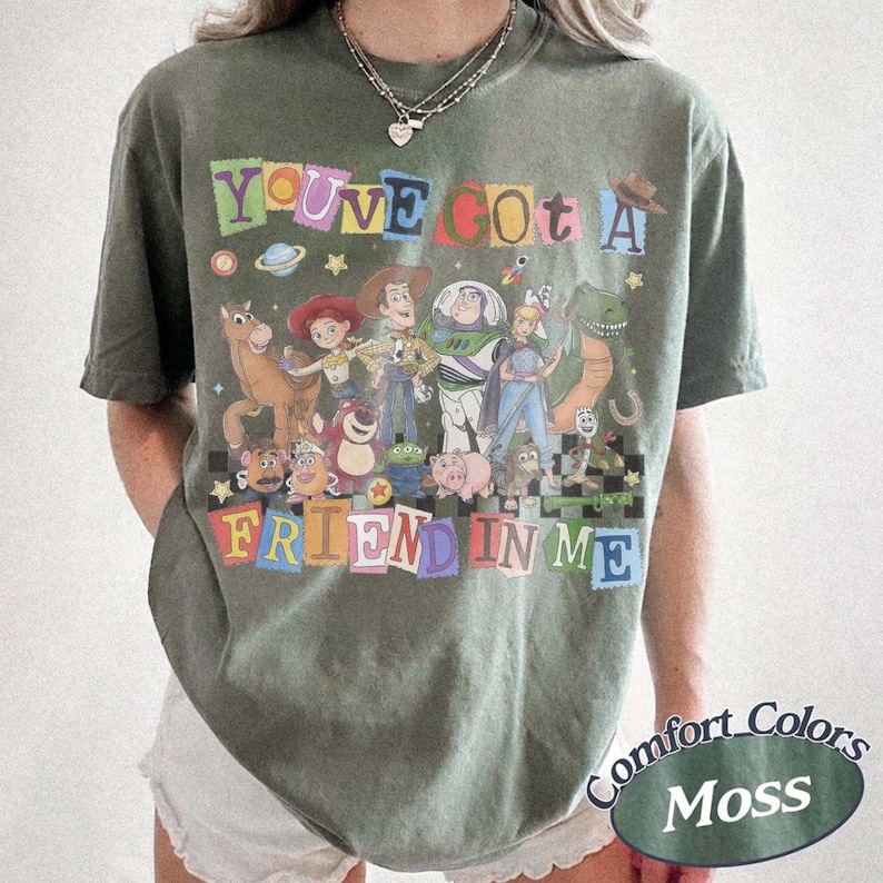 May include: A moss green short-sleeve t-shirt featuring a graphic print of various animated characters from the Toy Story franchise. The text "YOU'VE GOT A FRIEND IN ME" is printed in colorful, blocky letters above and below the character group.