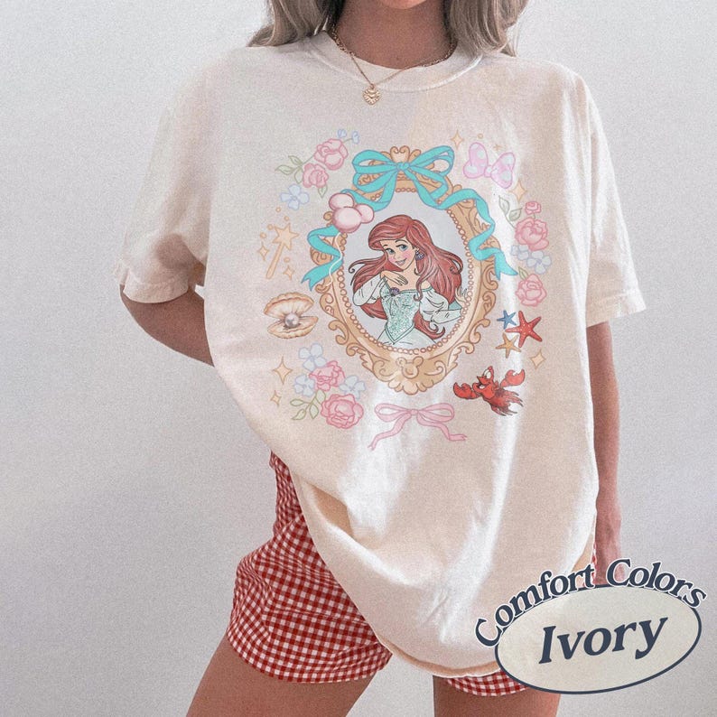 Puede incluir: Camiseta de color marfil con una ilustraci&oacute;n enmarcada de una mujer pelirroja, rodeada de flores rosas, lazos azules y un cangrejo rojo. La camiseta tiene las palabras "Comfort Colors" e "Ivory" impresas.