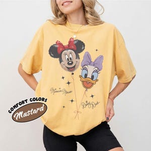 Pu&ograve; includere: Una t-shirt giallo senape con una stampa grafica di Minnie Mouse e Daisy Duck raffigurate come palloncini. Il design include il testo "Minnie Mouse" e "Daisy Duck" in corsivo, insieme a piccoli accenti a forma di stella. La t-shirt ha maniche corte.