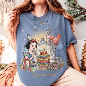 May include: Pale blue t-shirt with a Snow White design, a birthday cake, castle, and balloons. The number "2" and the text "Custom Name" are included. The shirt has a celebratory theme, perfect for a birthday.