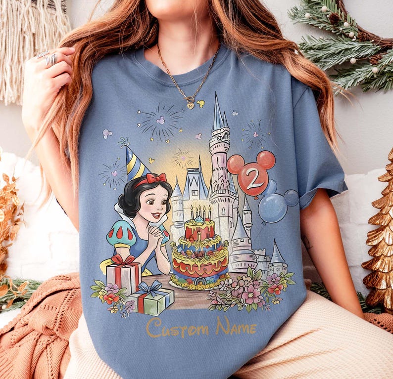 May include: Pale blue t-shirt with a Snow White design, a birthday cake, castle, and balloons. The number "2" and the text "Custom Name" are included. The shirt has a celebratory theme, perfect for a birthday.