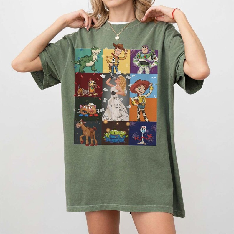 May include: An olive green short-sleeve t-shirt featuring a grid of nine colorful illustrations of various animated characters, including a green dinosaur, a cowboy, a space ranger, a dog, a blonde figure in a white dress, a cowgirl, potato figures, a horse, aliens, and a white spork character.