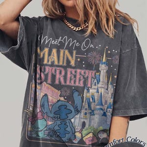 May include: Dark gray t-shirt with a graphic featuring Stitch from Lilo & Stitch, the text "Meet Me On Main Street", and a castle illustration. The shirt is labeled "Comfort Colors Pepper".