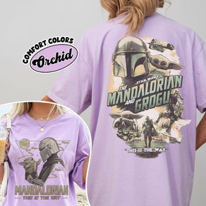 Op de afbeelding: Orchidee-kleurig T-shirt met afbeeldingen uit de serie "The Mandalorian". De voorkant toont de Mandalorian en Grogu, terwijl de achterkant een groter ontwerp heeft met de tekst "The Mandalorian and Grogu" en "This is the Way."