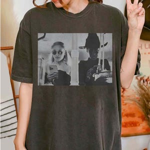May include: Dark gray t-shirt featuring a black and white image of two people holding phones. One person wears sunglasses and the other wears a hat. The image is a vintage style print.