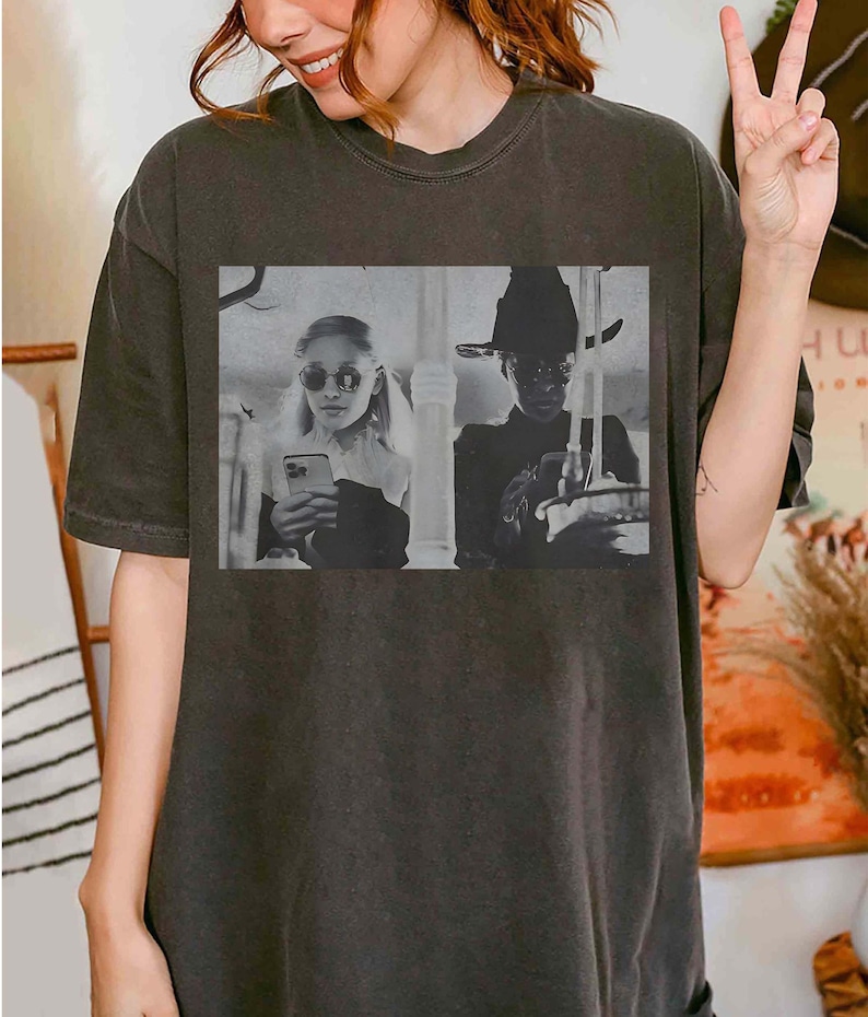May include: Dark gray t-shirt featuring a black and white image of two people holding phones. One person wears sunglasses and the other wears a hat. The image is a vintage style print.