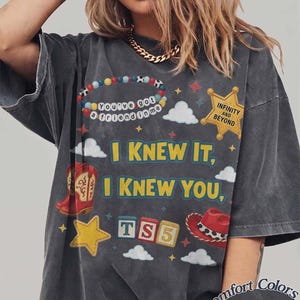 May include: A charcoal gray short-sleeve t-shirt featuring a graphic print with the text "I KNEW IT, I KNEW YOU," "You've got a friend in me," "INFINITY AND BEYOND," and "TS5." The design includes a beaded bracelet, a yellow star badge, clouds, red cowboy boots, a yellow star, and a red cowboy hat.