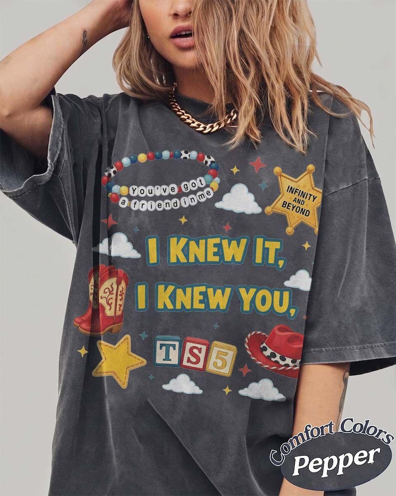 May include: A charcoal gray short-sleeve t-shirt featuring a graphic print with the text "I KNEW IT, I KNEW YOU," "You've got a friend in me," "INFINITY AND BEYOND," and "TS5." The design includes a beaded bracelet, a yellow star badge, clouds, red cowboy boots, a yellow star, and a red cowboy hat.