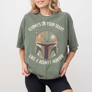 May include: An olive green t-shirt with a graphic of a helmet and the text "Karma's on your scent like a bounty hunter." The helmet design uses brown, green, and tan. The shirt has short sleeves and a loose fit.