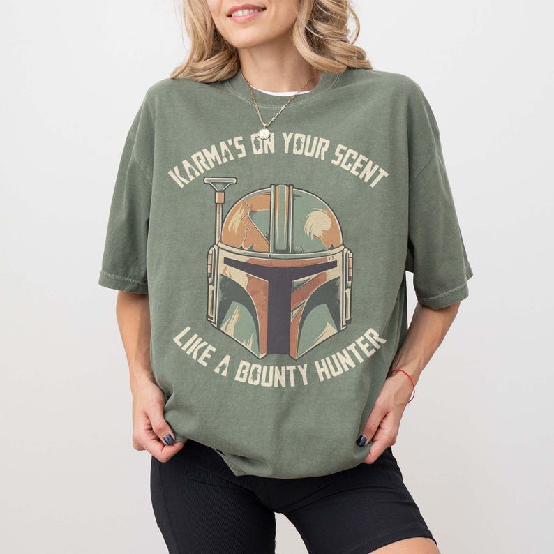 May include: An olive green t-shirt with a graphic of a helmet and the text "Karma's on your scent like a bounty hunter." The helmet design uses brown, green, and tan. The shirt has short sleeves and a loose fit.