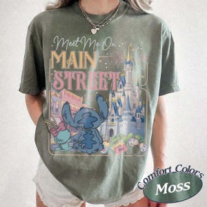 May include: Moss green t-shirt featuring a graphic of Stitch and Angel in front of a castle with the text "Meet Me On Main Street". The shirt also has the words "Comfort Colors" and "Moss" printed on it.