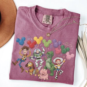 May include: A mauve short-sleeve t-shirt featuring a graphic print of Toy Story characters including Woody, Buzz Lightyear, Rex, Jessie, Slinky Dog, and Hamm with colorful Mickey Mouse-shaped balloons. The shirt has a Comfort Colors brand label.