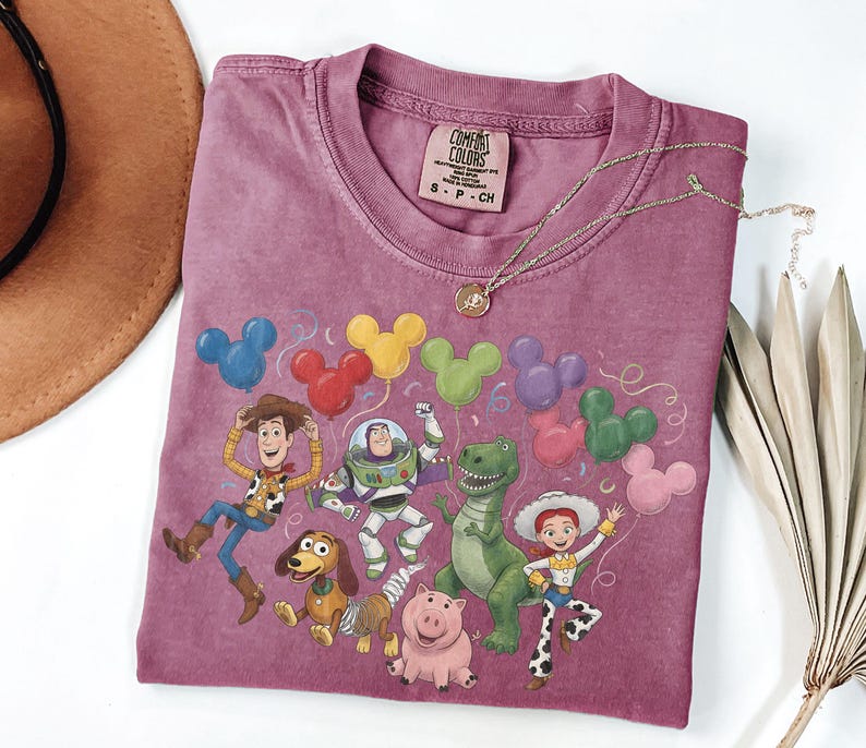 May include: A mauve short-sleeve t-shirt featuring a graphic print of Toy Story characters including Woody, Buzz Lightyear, Rex, Jessie, Slinky Dog, and Hamm with colorful Mickey Mouse-shaped balloons. The shirt has a Comfort Colors brand label.
