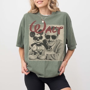 Pu&ograve; includere: Una t-shirt a maniche corte color verde oliva con una stampa grafica di un uomo e un topo dei cartoni animati, entrambi con occhiali da sole. Il testo "WALT" &egrave; stampato in un carattere rosso consumato sopra l'immagine in bianco e nero.