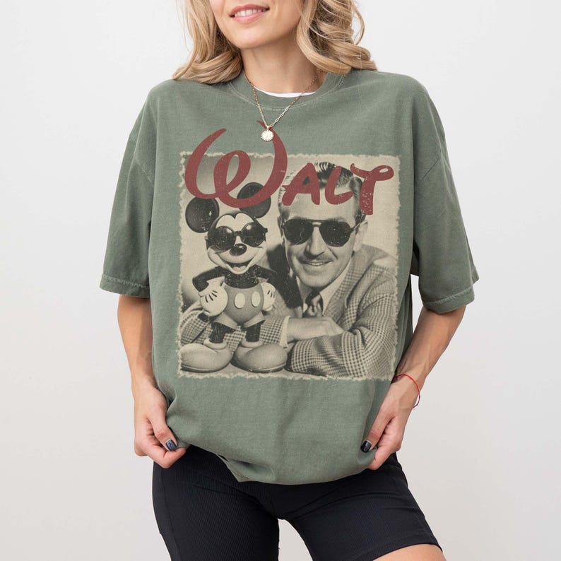 Pu&ograve; includere: Una t-shirt a maniche corte color verde oliva con una stampa grafica di un uomo e un topo dei cartoni animati, entrambi con occhiali da sole. Il testo "WALT" &egrave; stampato in un carattere rosso consumato sopra l'immagine in bianco e nero.