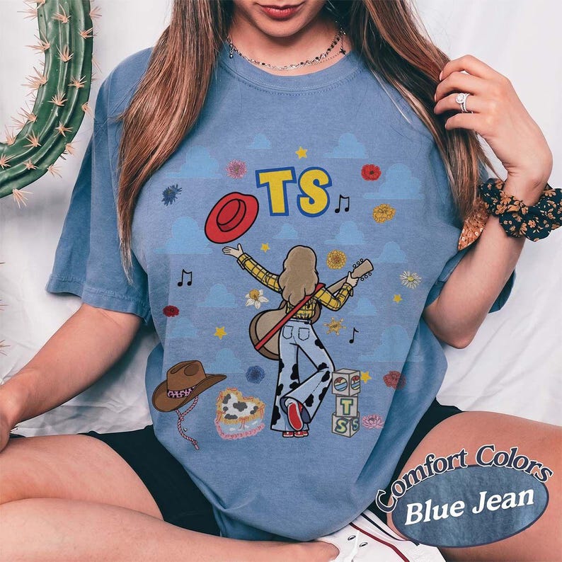 May include: A blue jean colored t-shirt featuring a graphic print of a person with a guitar, a red hat, and cow-print pants. The design includes the text TS, musical notes, flowers, and a stack of blocks. The background has a light blue cloud pattern.
